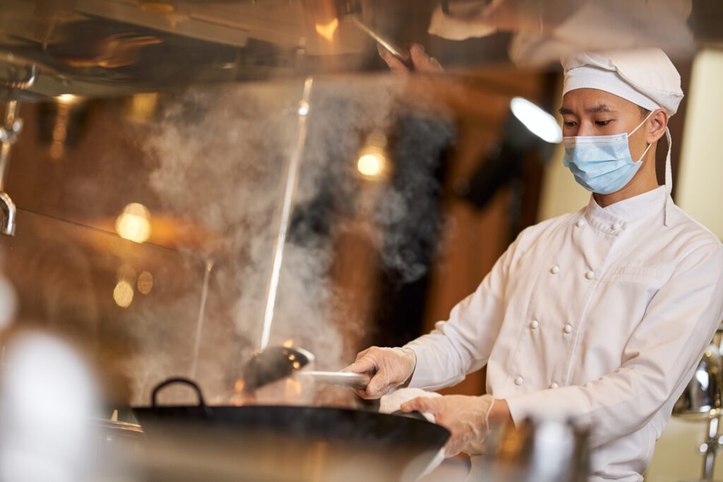 Hard-working Asian chef in protective mask and uniform standing near stove while stirring in steamy wok