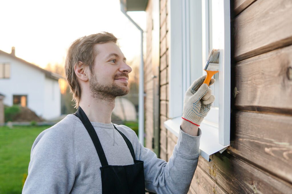 Young smiling man in apron painting window of his house with paint brush.
